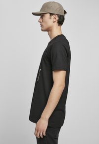 T-shirt en coton noir à manches courtes, avec un motif crème, associée à une casquette beige. Le design est simple et décontracté.