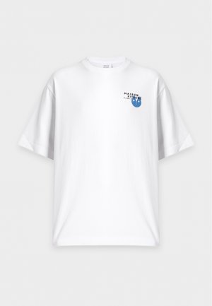 Ung mand står iført en hvid T-shirt med et lille blåt logo, falmede blå jeans og hvide sneakers mod en ensfarvet baggrund.