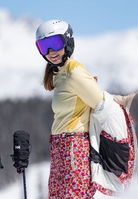 Ski oblečení se žlutým dlouhým rukávem jako základní vrstvou, lyžařskými kalhotami s květinovým vzorem a bílou vycpanou bundou. Součástí jsou také černé lyžařské rukavice a brýle.