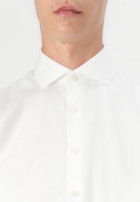 Camicia bianca con colletto classico, caratterizzata da una texture liscia e una leggera lucentezza. Sei bottoni rotondi al centro.