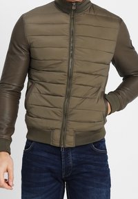 Man med olivgrön vadderad jacka med läderärmar och mörkblå jeans, en hand i fickan och dragkedjan stängd.