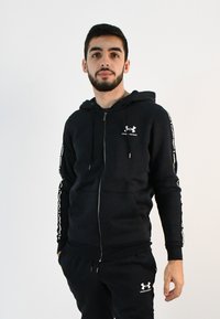 Sudadera con cremallera negra de material suave, con un logo en el pecho y detalles blancos a lo largo de las mangas. Combinada con pantalones a juego.