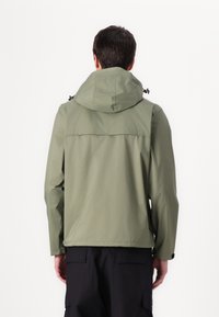 Ελαιοπράσινο αδιάβροχο μπουφάν με κουκούλα, που διαθέτει ραφή στην πλάτη και ρυθμιζόμενο �κορδόνι, συνδυασμένο με μαύρο cargo παντελόνι.