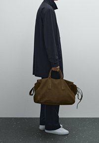 Braune Wildleder-Duffle-Tasche mit doppelten Obergriffen, geschwungenen Kanten und Metallbeschlägen. Die Tasche wird von einer Person getragen, die einen marineblauen Mantel trägt.