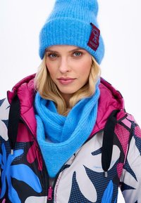 Barevná prošívaná bunda s růžovými akcenty a abstraktními květinovými vzory. Na sobě má jasně modrou pletenou čepici a odpovídající šálu. Hladká textura.