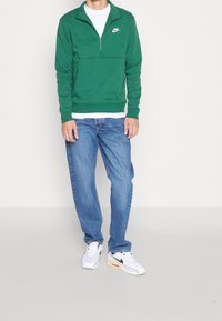 Zelená mikina na zip s bílým logem Nike, doplněná modrými džínami a bílými teniskami s vícebarevnými akcenty.