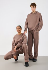 Brązowa bluza z kapturem i dopasowane spodnie dresowe z miękkiego, teksturowanego materiału. Charakteryzuje się luźnym krojem i ściągaczami na rękawach. Całość dopełniają czarne sneakersy.