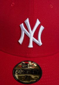Crvena bejzbol kapa s bijelim izvezenim NY logom na prednjoj strani. Ima ravni vizir i zlatnu naljepnicu s detaljima o veličini na viziru.
