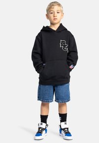 Sudadera negra con un logo blanco, pantalones cortos de mezclilla, zapatillas negras y azules, y calcetines negros. Estilo casual, corte relajado con un bolsillo frontal.
