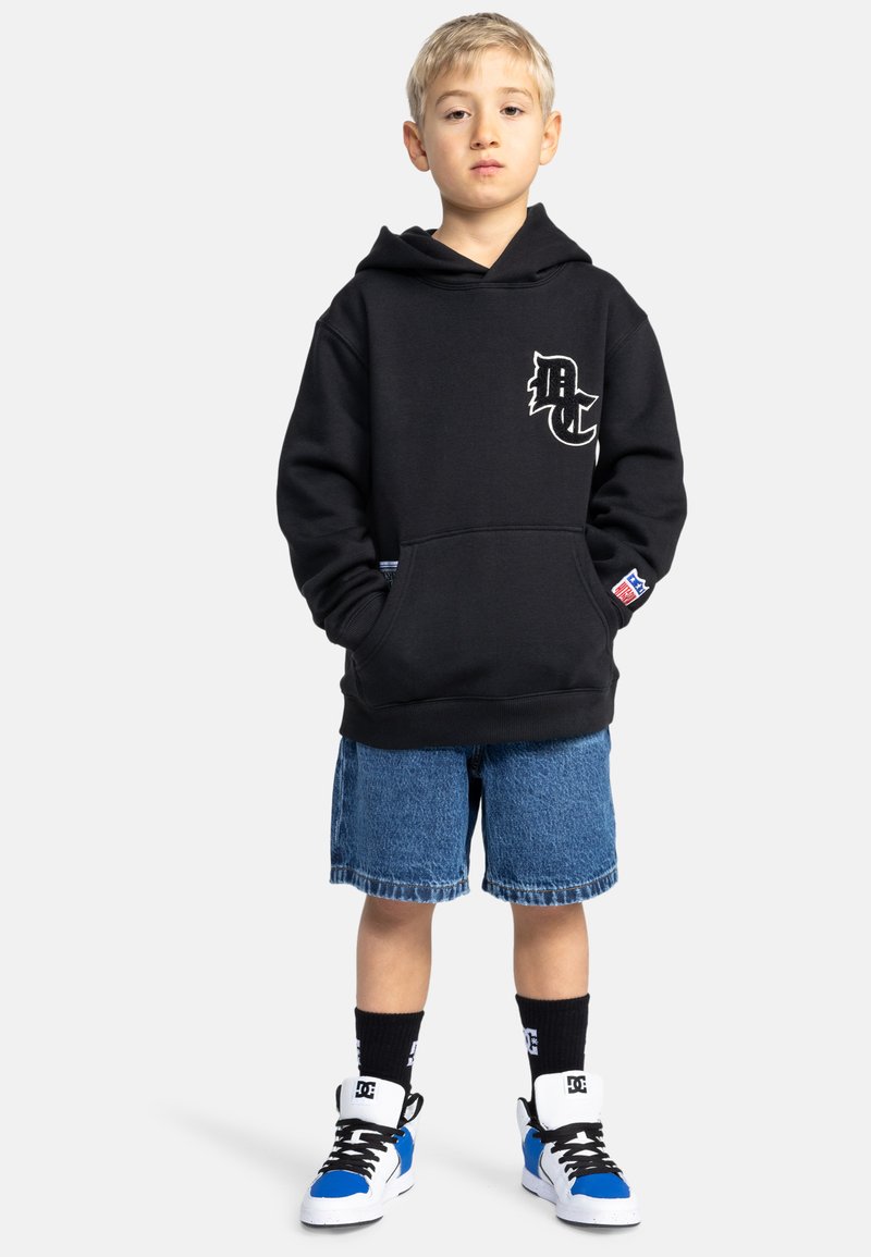 Sudadera negra con un logo blanco, pantalones cortos de mezclilla, zapatillas negras y azules, y calcetines negros. Estilo casual, corte relajado con un bolsillo frontal.