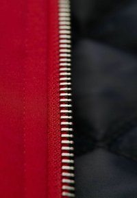Roter Stoff mit einer glatten Textur neben einem silbernen Reißverschluss. Die Reißverschlusszähne sind eng beieinander angeordnet, im Kontrast zu einem dunklen Hintergrund.