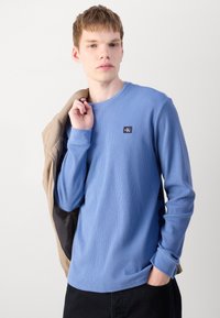 Lyseblå langermet ribbestrikket bomulls T-skjorte med rund hals og merket lapp på brystet. Modellen holder en beige jakke.
