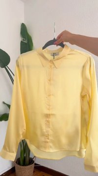 Main tenant un cintre noir avec une chemise jaune à manches longues boutonnée devant un mur blanc et une plante en pot.