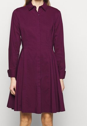Frau trägt ein langärmliges, geknöpftes Kleid mit Kragen in dunklem Lila, mit tailliertem Schnitt und ausgestelltem Rock, steht vor einem schlichten Hintergrund.