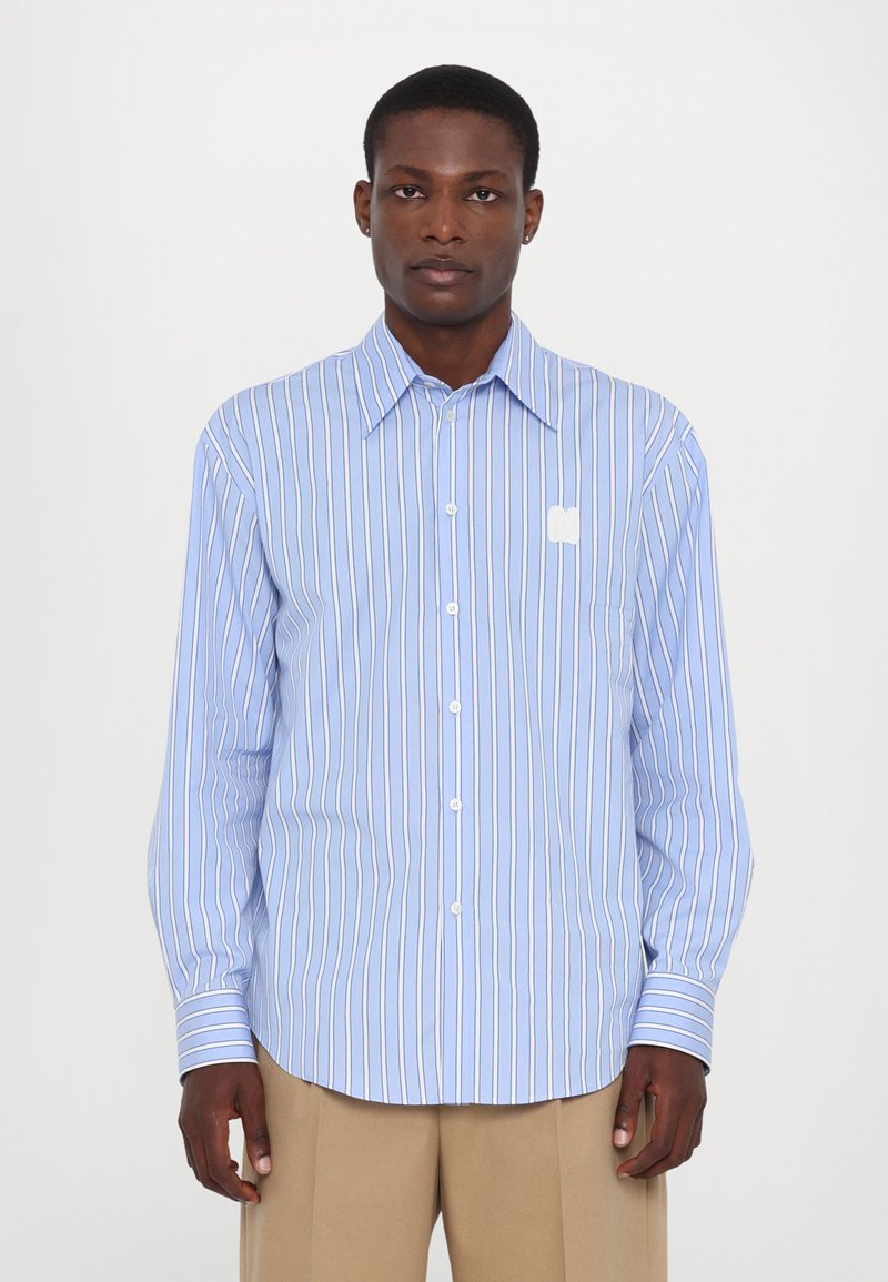 Camisa de manga larga azul claro con rayas verticales blancas y gris claro, con botones en la parte delantera y un pequeño logo blanco en el bolsillo del pecho izquierdo.
