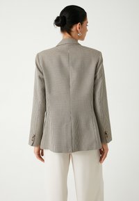 Blazer a motivo pied-de-poule in beige e nero, con una vestibilità sartoriale, rever a intaglio e polsini con bottoni. Indossato sopra pantaloni color crema.