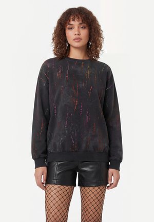 Kvinde med krøllet hår iført en sort sweatshirt med flerfarvede striber, sorte lædershorts og netstrømpebukser, stående mod en hvid baggrund.
