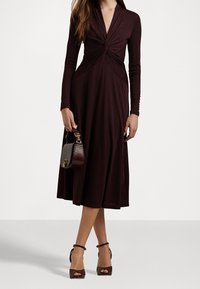 Bordeaux långärmad klänning med tvist fram, flödande kjol och textilstruktur. Kombinerad med en liten mörk handväska och klacksandaler.