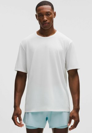 Joven hombre con una camiseta blanca lisa de manga corta y pantalones cortos deportivos azul claro, de pie frente a un fondo claro y liso.