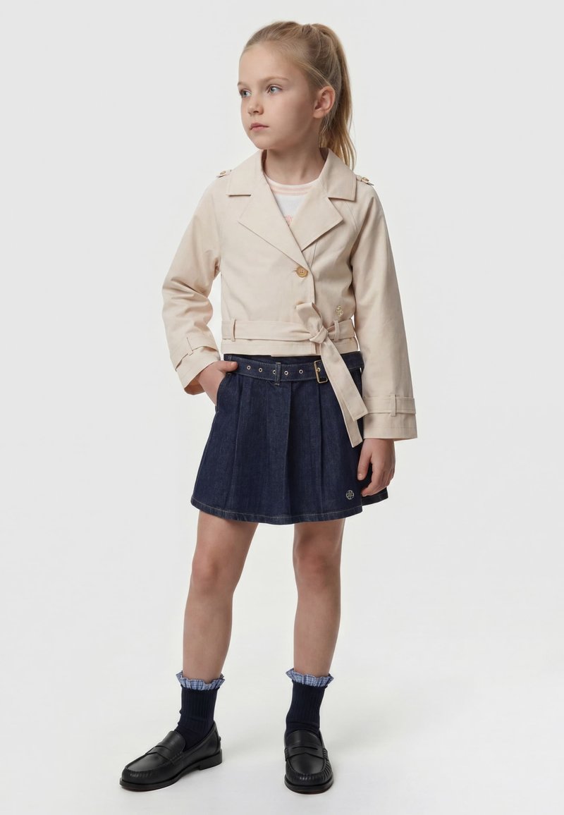 Jeune fille aux cheveux blonds portant une veste beige ceinturée, une jupe plissée foncée, des chaussures noires et des chaussettes à volants bleu marine, debout les mains dans les poches.