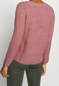 Gebreide trui in stoffige roze met een gestructureerd patroon, ronde halslijn en lange mouwen. Gecombineerd met donkergroene leggings.