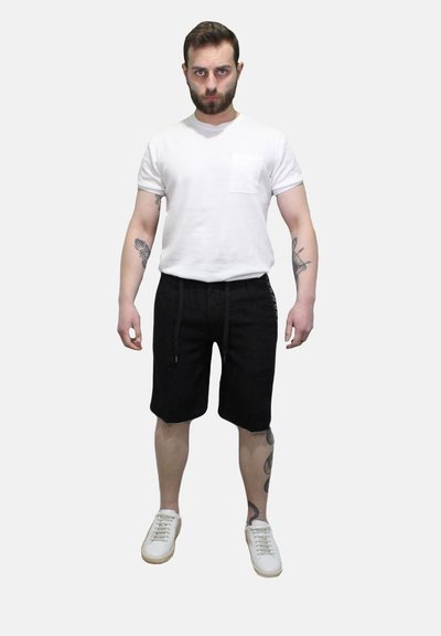 T-shirt bianca a girocollo con taschino, pantaloni corti neri con coulisse e sneakers bianche. Presenta tatuaggi sulle braccia e uno sfondo neutro.