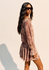 Lila gemustertes off-shoulder Kleid mit langen Ärmeln und gerafftem Saum. Weiches Material, fließendes Design und sanfter Schimmer. Getragen mit dunkler Sonnenbrille.
