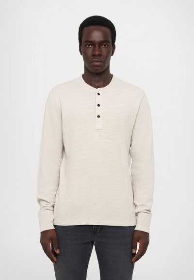 Chemise henley à manches longues en beige clair, fabriquée en tissu texturé avec un col rond, trois boutons noirs et des poignets ajustés.