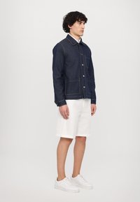 Jeanjakke i mørkeblått med synlige sømmer, knappestolpe og lapper på lommer, paret med hvite shorts og sneakers.