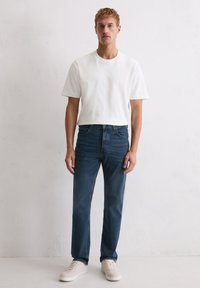 Homme se tenant contre un mur blanc, portant une chemise blanche à manches courtes, un jean bleu foncé et des baskets beiges, faisant face à l'avant avec les mains détendues le long du corps.