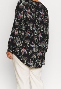 Person wearing black blouse with red, blue, and white paisley pattern and white pants, shown from the back against a plain background.