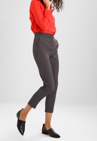 Pantaloni neri a fantasia con vita alta, gamba affusolata e un design geometrico sottile. Abbinati a una blusa rossa maniche lunghe e scarpe nere.