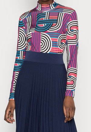 Femme portant un top à manches longues à motif géométrique multicolore avec un col montant et une jupe plissée bleu marine.