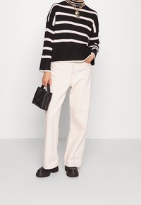 Osoba ubrana w czarno-biały sweter w paski, białe szerokie spodnie, czarne masywne buty, złoty naszyjnik, trzymająca małą czarną torebkę.