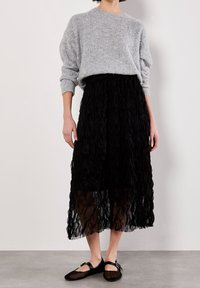 Personne portant un pull en maille gris clair rentré dans une jupe midi texturée noire avec des ballerines Mary Jane noires, debout sur un sol gris.