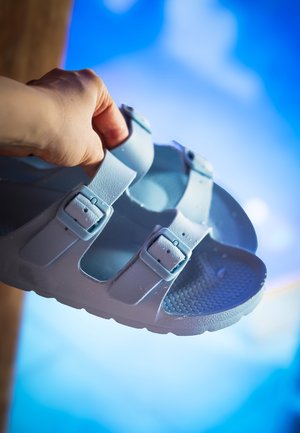 Hand holding a light blue sandal with two adjustable buckle straps against a blurred blue sky background.