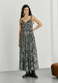 Sieviete stāv telpās, valkājot melnu un baltu rakstainu maksi kleitu ar plānām plecu siksnām un melnas potīšu zābaciņus, roka uz gurniem.