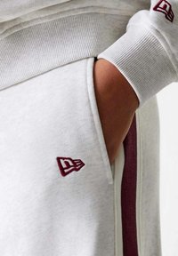 Weißer Sweatshirt mit gerippten Bündchen, versehen mit einem burgunderfarbenen Logo am Ärmel. Enthält eine Tasche mit einem Streifendetail an der Seite.