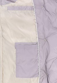 Vadderad jacka i ljus lavendel med en beige insida. Har en ficka och knappdetalj, slät textur och kontrasterande kant.