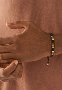 Flätad armband med brun snodd, tre guldfärgade metallaccenter och en justerbar stängning. Buren på en hand mot en texturerad bakgrund.