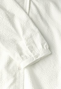 Camicia bianca texturizzata con polsini a bottone, tessuto intrecciato con motivo a pois e un aspetto leggermente stropicciato.