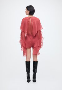 Γυναίκα που φοράει διάφανο, ρέον κόκκινο ολόσωμο φόρεμα με στρωτές μανίκες και μαύρα ψηλά μποτάκια μέχρι το γόνατο, στέκεται γυρισμένη με την πλάτη σε λευκό φόντο.