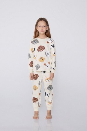 Jeune fille debout, pieds nus, portant un pyjama blanc orné de visages de chiens colorés sur un fond clair uni.