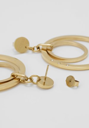 Boucles d'oreilles créoles ton or avec un pendentif circulaire et de petites pierres claires, une posée à plat montrant la tige et le fermoir papillon sur fond blanc.
