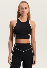 Čierny atletický crop top s bielymi detailmi; má vysoký golier a priliehavý strih. Kombinovaný s čiernymi legínami s vysokým pásom.