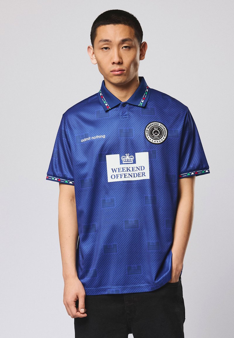 Jeune homme portant un polo bleu "Weekend Offender" avec col et bordure à motifs, debout avec une expression neutre sur un fond uni.