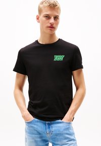 T-shirt negru din bumbac cu un logo grafic verde aprins pe partea stângă a pieptului, guler rotund și mâneci scurte. Purtați cu blugi de culoare albastru deschis.