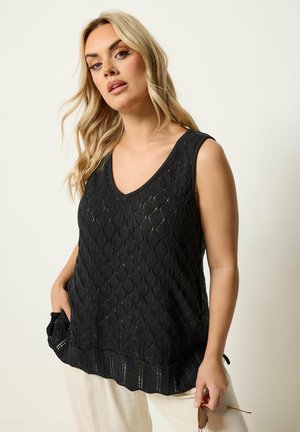 Blonde Frau trägt ein schwarzes ärmelloses Stricktop und helle Hosen, hält eine Sonnenbrille in einer Hand, vor einfarbigem Hintergrund.