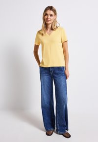 Geel kort mouwtop met een V-hals ontwerp, gecombineerd met brede blauwe denim jeans en zwarte mesh schoenen. Gladde stof met een aansluitende silhoutte.