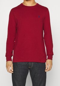 Röd långärmad t-shirt i bomull med rund hals och åtsittande ärmar. Har en liten navy-logotyp på bröstet. Kombinerad med mörka jeans.